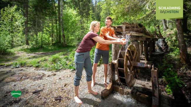 Zwei Wanderer erfrischen sich nach einer anstrengenden Tour in einem Bach. Neben ihnen steht ein riesiges, rustikales Holzrad, das idyllisch vom klaren Wasser umspült wird.