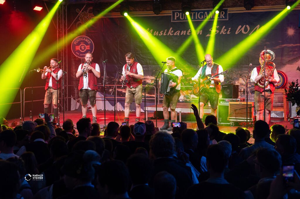 Sechs Künstler stehen in traditionellen Lederhosen auf einer Bühne und spielen gemeinsam ihre Instrumente vor Publikum.