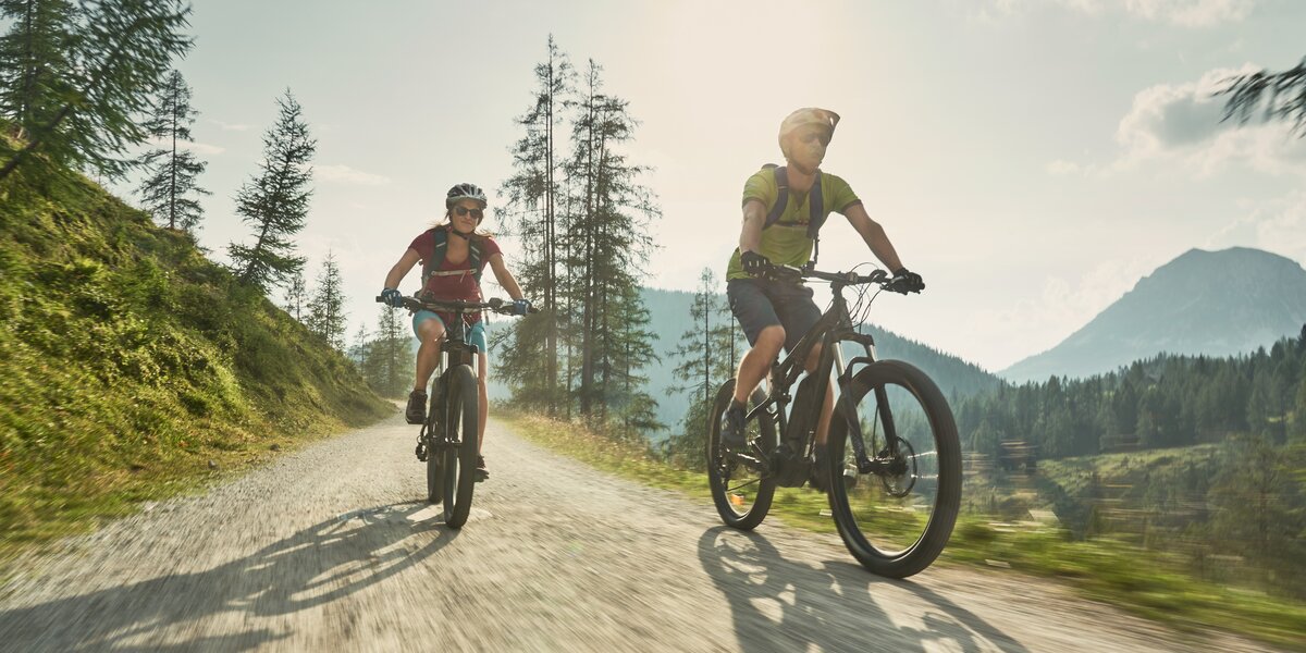 Zwei Personen sitzen auf einem Fahrrad und rasen gemeinsam einen schmalen Schotterweg entlang.