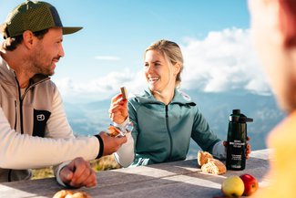 Wanderpaar genießt bei einer Pause mit Bergpanorama eine Jause an einem Holztisch.