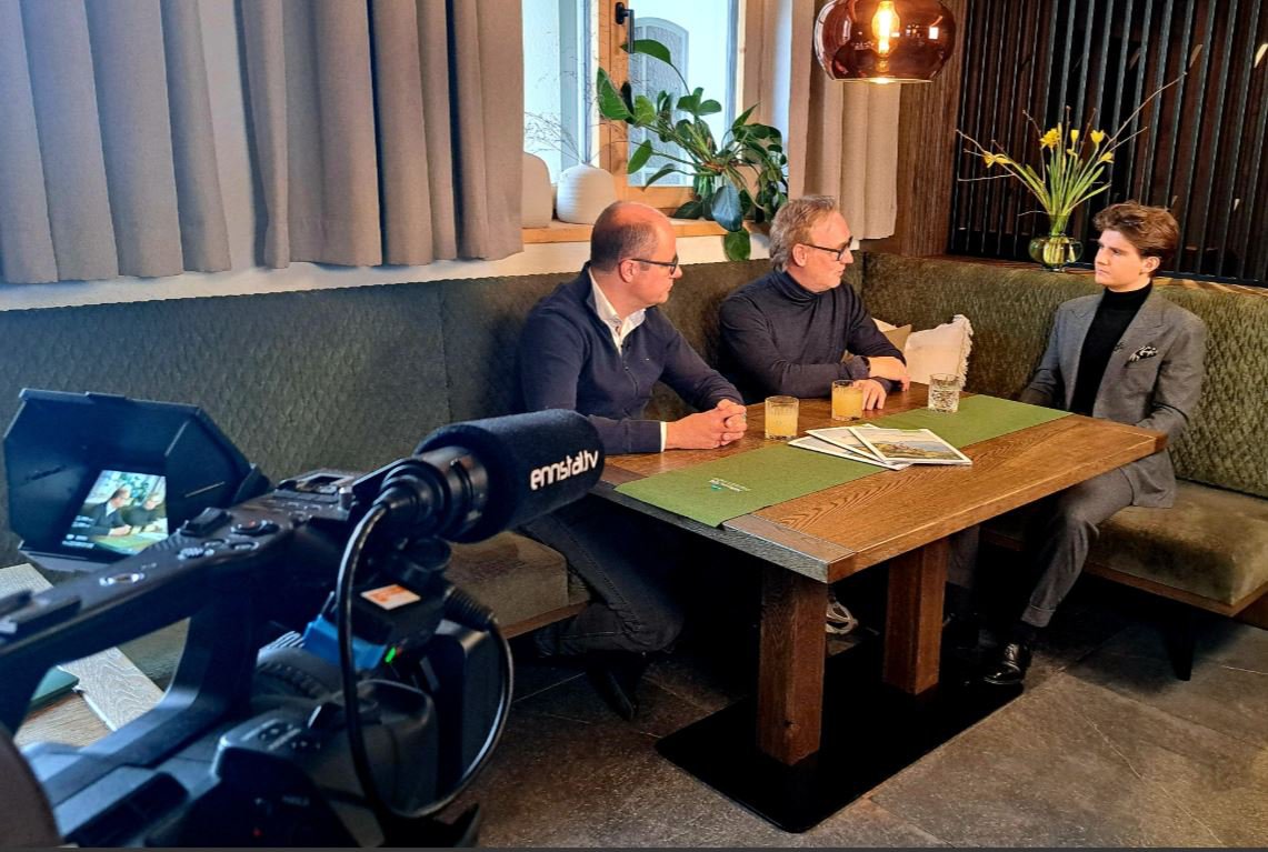 Drei Personen sitzen an einem Tisch und sind in ein intensives Gespräch vertieft und das Gespräch wird von einer Kamera aufgenommen.