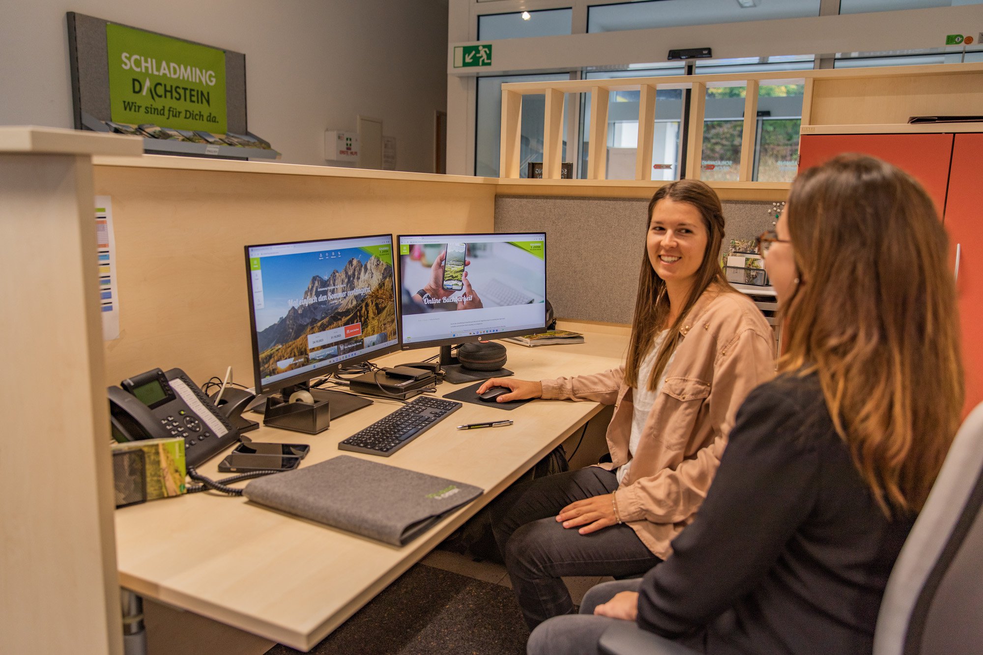 Zwei Frauen sitzen nebeneinander, und eine zeigt der anderen etwas auf dem Computerbildschirm. Beide lächeln sich an und sind im Gespräch vertieft.