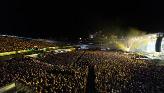 Ein großes Open-Air-Konzert bei Nacht mit einer riesigen Bühne und tausenden begeisterten Zuschauern.