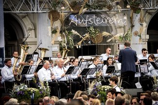 Ein großes Orchester spielt gemeinsam auf einer Konzertbühne, während ein charismatischer Dirigent mit erhobenen Armen konzentriert den Takt vorgibt.