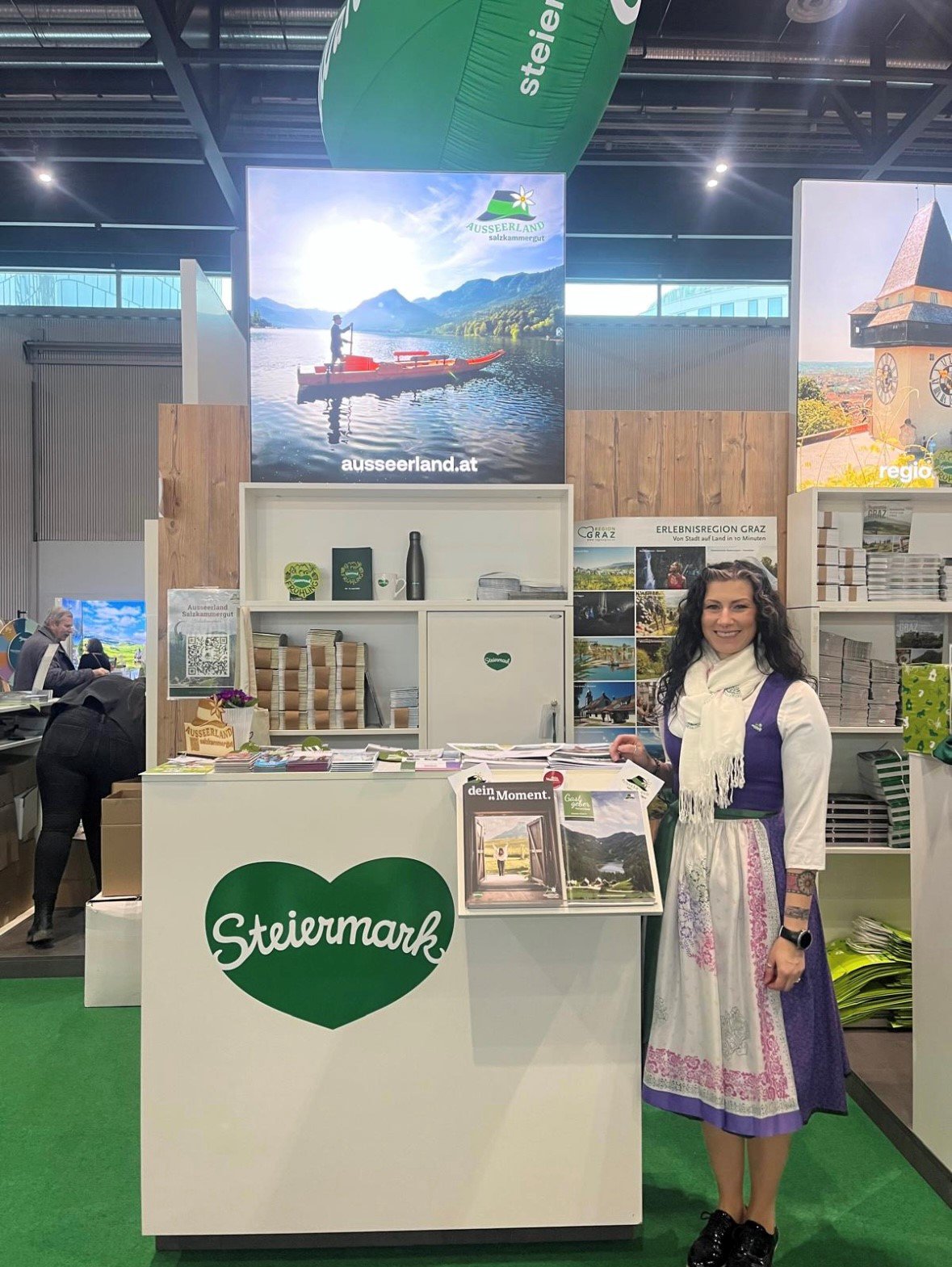 Ausseerland Stand mit Plättenbild im Hintergrund. Neben den Pult steht Nathalie Schröttenhamer im Dirndl.