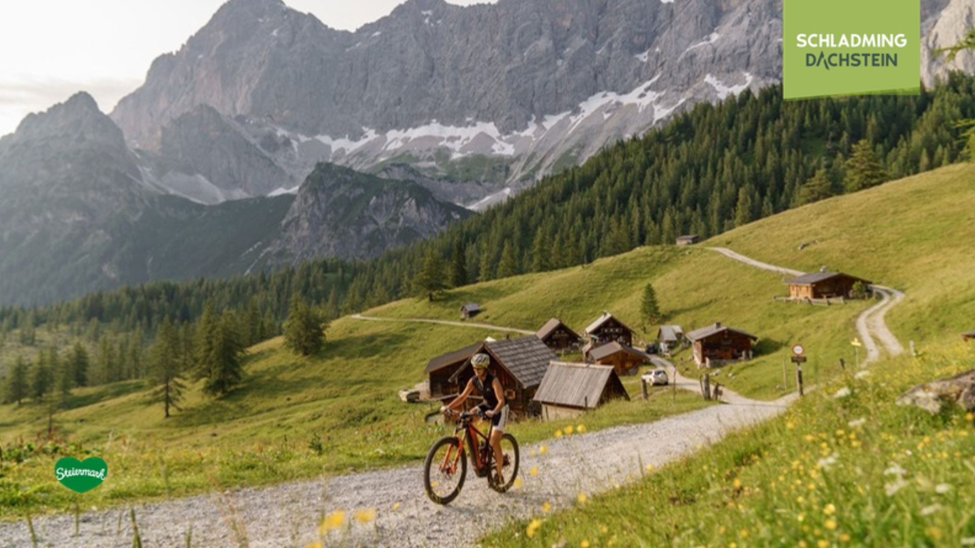 Eine Frau fährt mit dem Mountainbike auf einem Schotterweg durch die Berge. Im Hintergrund erstrecken sich idyllische Almen und das beeindruckende Dachsteinmassiv.