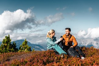 Zwei Personen sitzen inmitten einer herbstlichen Wiesenlandschaft, während sich im Hintergrund ein atemberaubender Blick auf die imposante Bergkulisse eröffnet.
