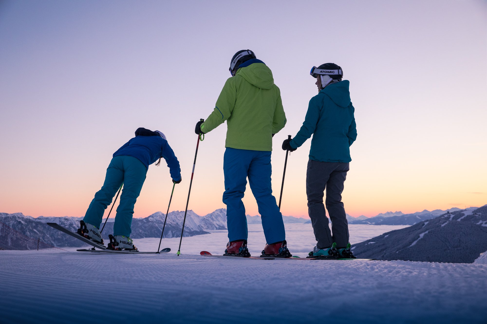 Drei Personen stehen vor einem Pistenabhang, im Hintergrund zeigt sich ein verschneites Bergpanorama in den Morgenstunden mit schöner Nebelschicht.