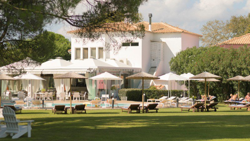 Blick auf den Pool und den Garten des Mandraki Village mit Liegestühlen, üppiger Begrünung und entspannter Atmosphäre | © Mandraki Village