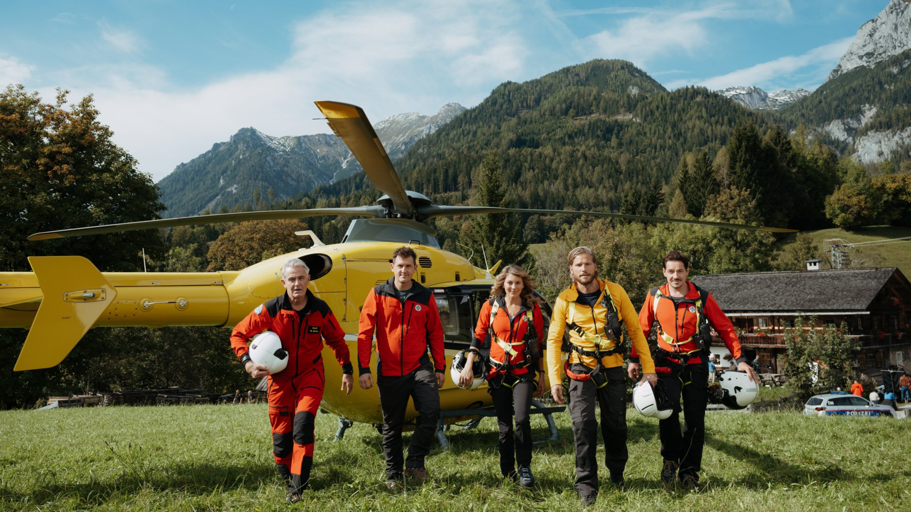 Die TV-Serie Die Bergretter mit den Bergretter-Schauspielern im Vordergrund und einem gelben Helikopter im Hintergrund.