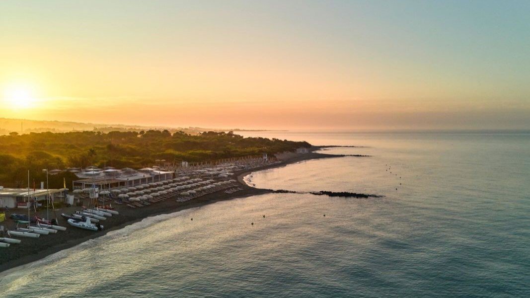 Luftaufnahme einer ruhigen Küstenlandschaft bei Sonnenaufgang mit sanftem Wellengang, einem Strand mit Sonnenschirmen und kleinen Booten am Ufer | © ROBINSON
