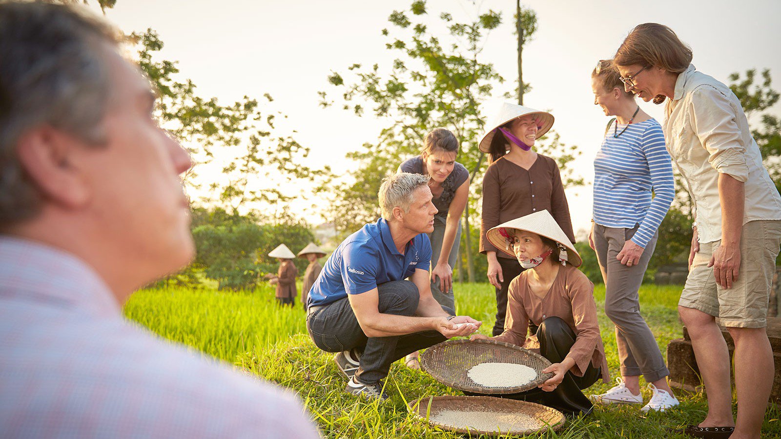 Reisbäuerin in Vietnam zeigt ein Sieb mit Reis, während die Studiosus-Reiseleitung die Verarbeitung des Reises einer Besuchergruppe erklärt | © Studiosus