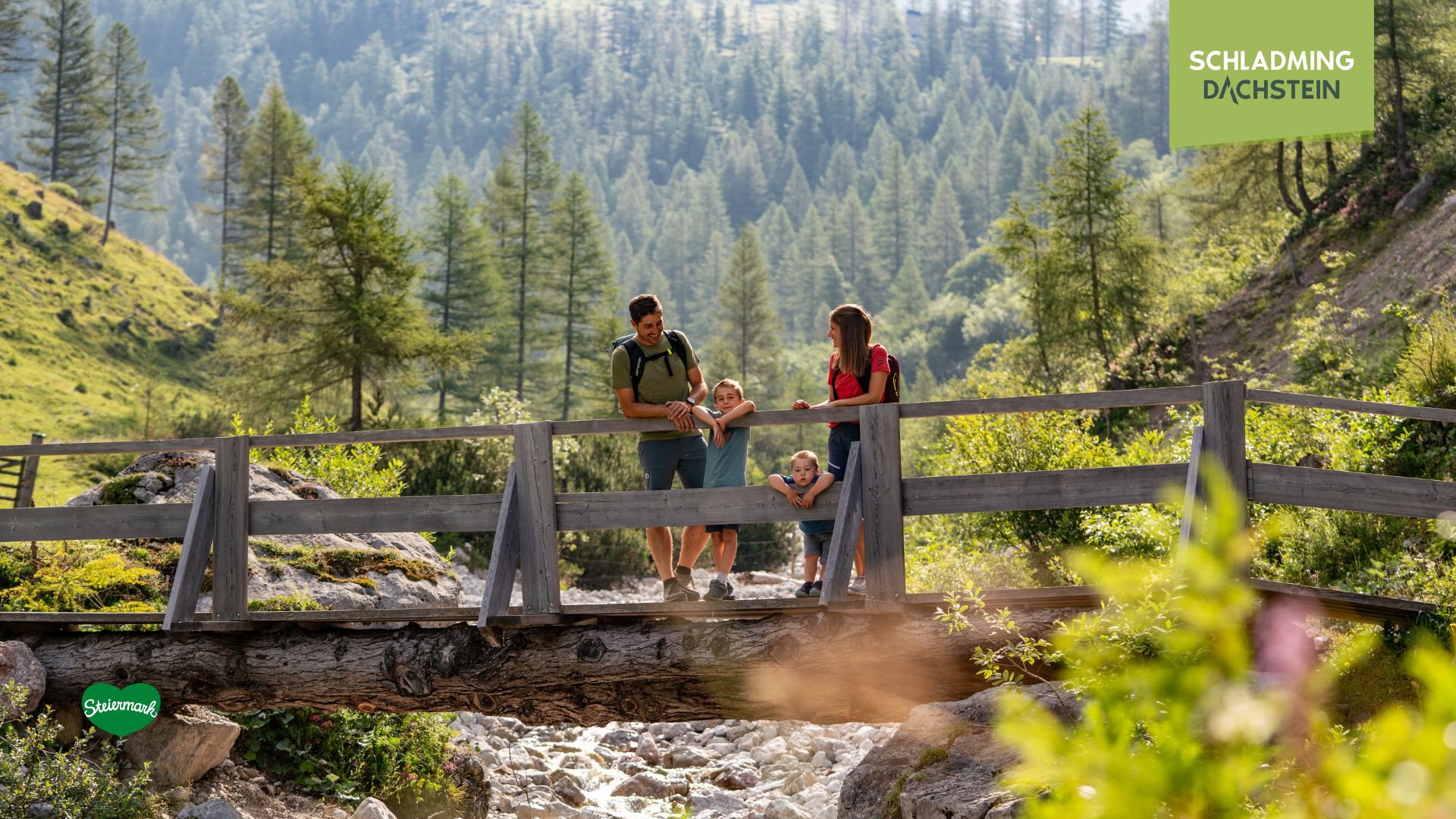 Eine Familie mit zwei Kindern genießt einen sonnigen Wandertag und steht gemeinsam auf einer Holzbrücke, umgeben von idyllischer Natur.