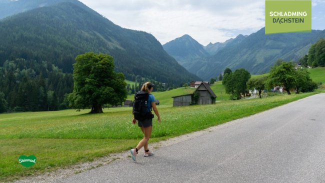 Eine Wanderin spaziert auf einem asphaltierten Weg, während sich im Hintergrund die imposanten Schladminger Tauern erheben.