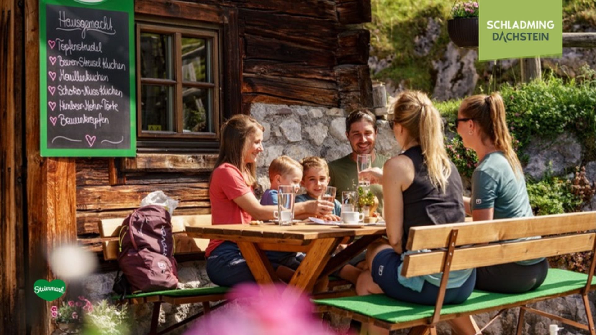 Eine fröhliche Familie sitzt an einem rustikalen Holztisch vor einer traditionellen Alpenalm in den Bergen.