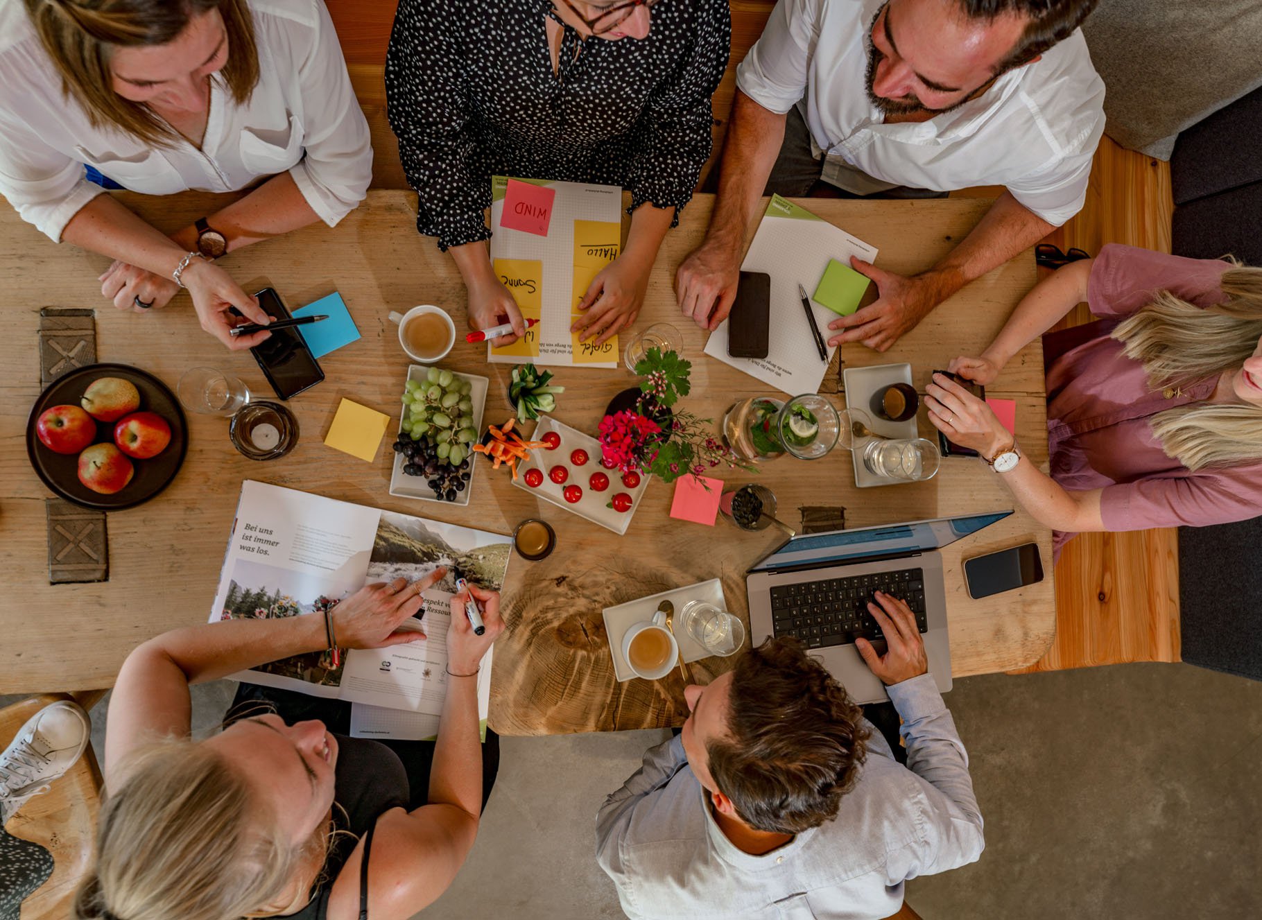 Sechs Personen sitzen gemeinsam an einem Tisch, auf dem verteilt Papiere, Laptops und Notizblöcke liegen, und besprechen miteinander ein Thema.