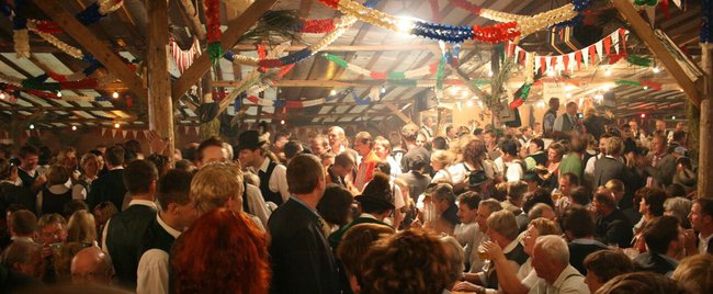 Das Holzstangenzelt der Freiwilligen Feuerwehr Altaussee von innen gefüllt von Menschen in Tracht und einer tobenden Stimmung. Girlanden und Lichter hängen von der Decke herunter. 