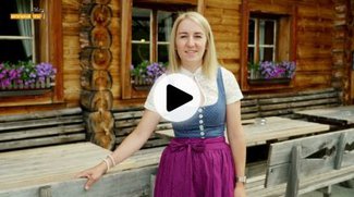 Eine Dame in einem traditionellen Dirndl steht vor einer gemütlichen Holzhütte in den Bergen.