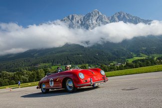 Ein rotes Auto fährt auf einer asphaltierten Straße. Im Hintergrund sieht man eine atemberaubende Bergkulisse.